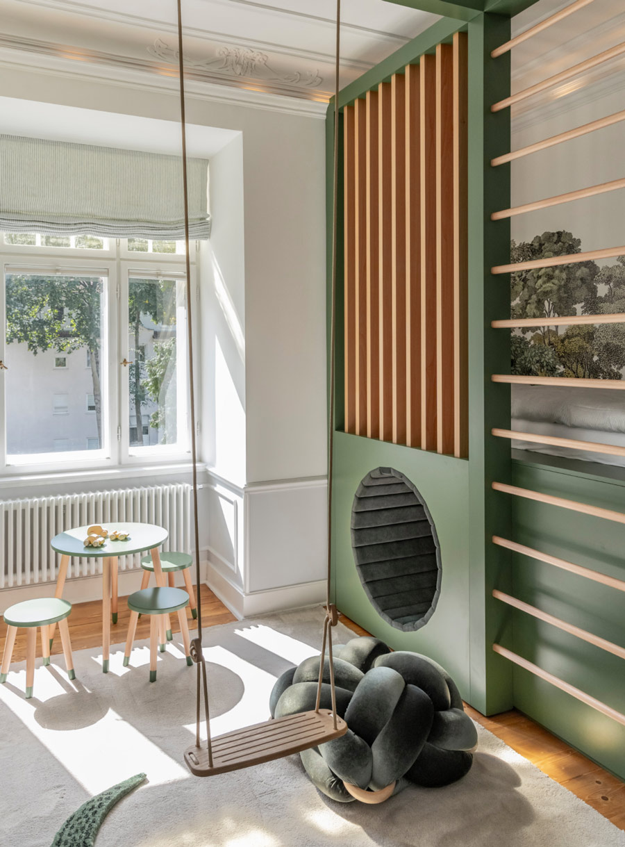 Zu sehen ist hier ein lichtdurchflutetes Spielzimmer mit integrierter Kletterwand, Holzschaukel, maßgefertigten dunkelgrünen Einbauten und heller Fensterfront im klassischen Altbau.